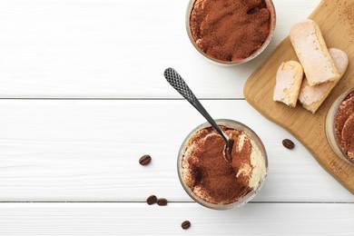 Photo of Delicious tiramisu in glasses, spoon, coffee beans and cookies on white wooden table, top view. Space for text