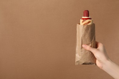 Photo of Woman holding paper bag with delicious french hot dog on brown background, closeup. Space for text