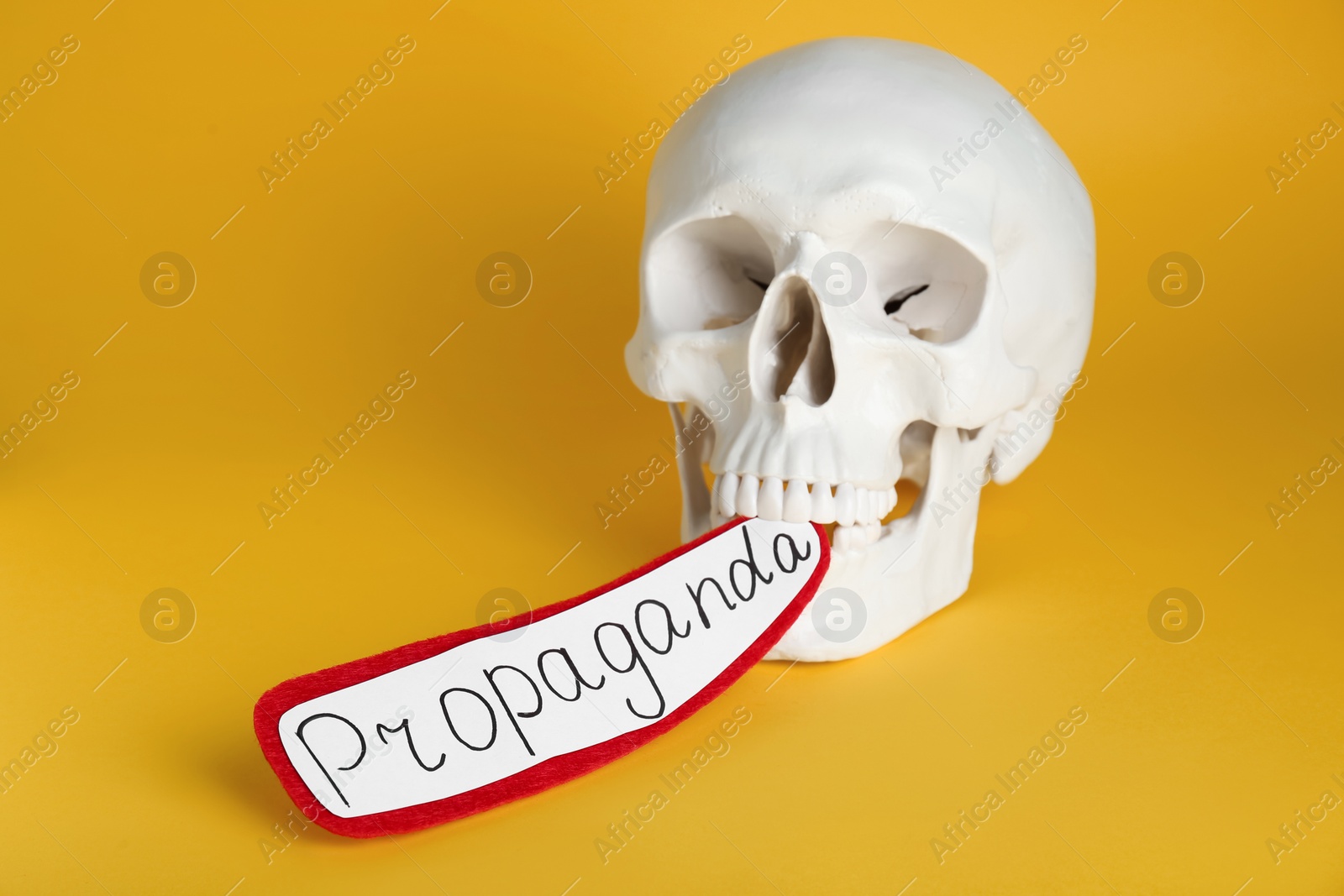 Photo of Information warfare concept. Human skull and paper card with word Propaganda in teeth on yellow background