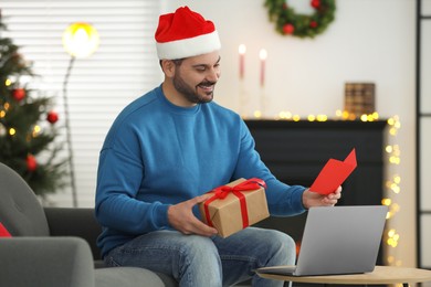 Photo of Celebrating Christmas online with exchanged by mail presents. Smiling man in Santa hat with greeting card and gift box during video call on laptop at home