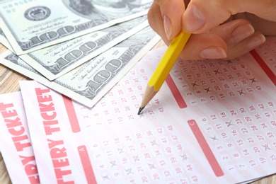 Photo of Woman filling out lottery tickets with pencil and money on wooden table, closeup. Space for text