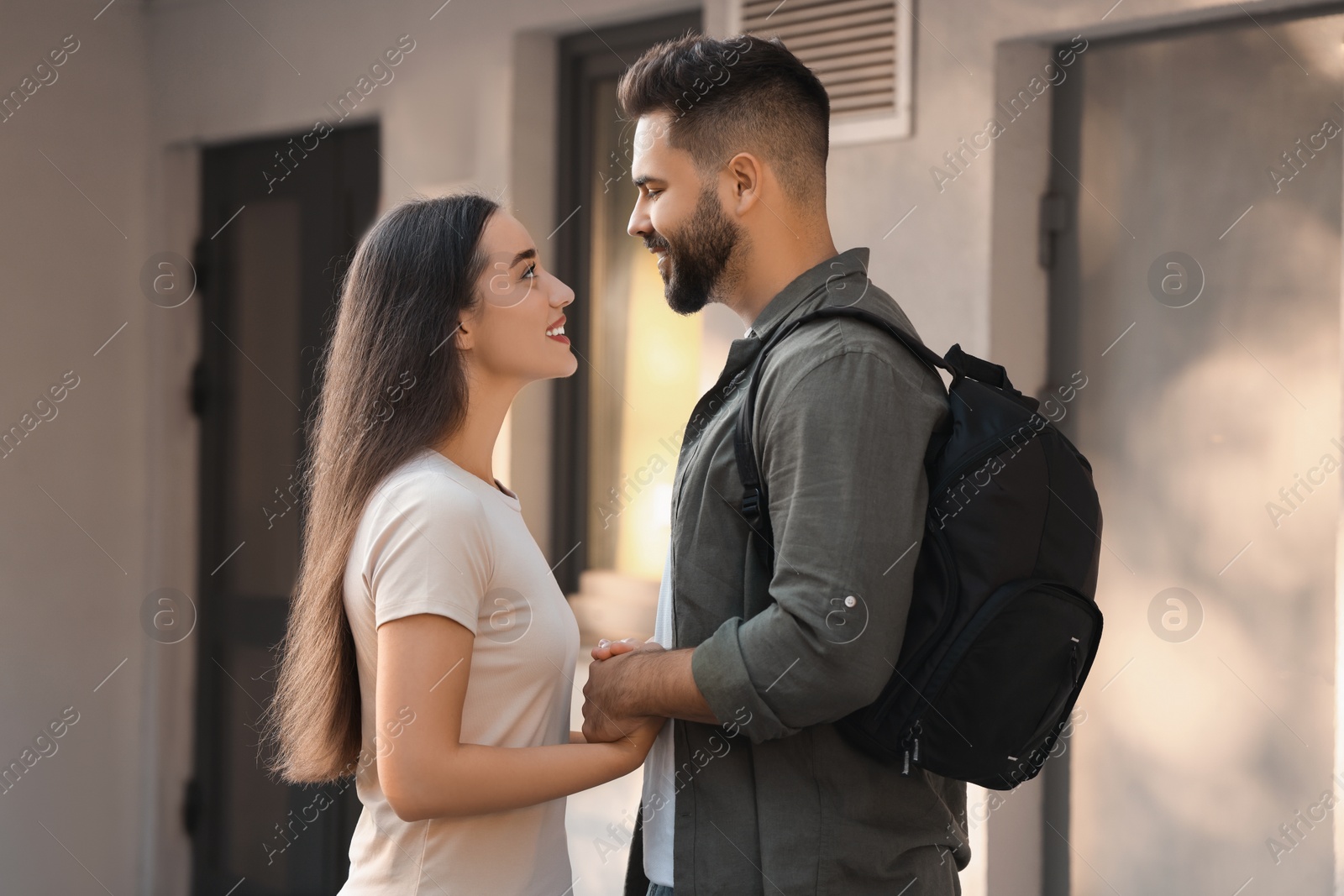 Photo of Long-distance relationship. Man with backpack holding hands with his girlfriend near house entrance outdoors