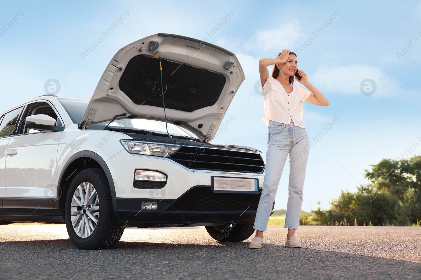 Photo of Stressed young woman talking on phone near broken car on roadside, low angle view