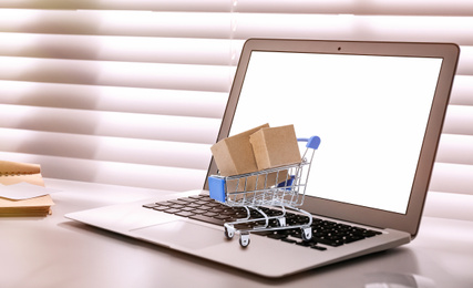 Image of Online shopping. Small cart with boxes on laptop indoors