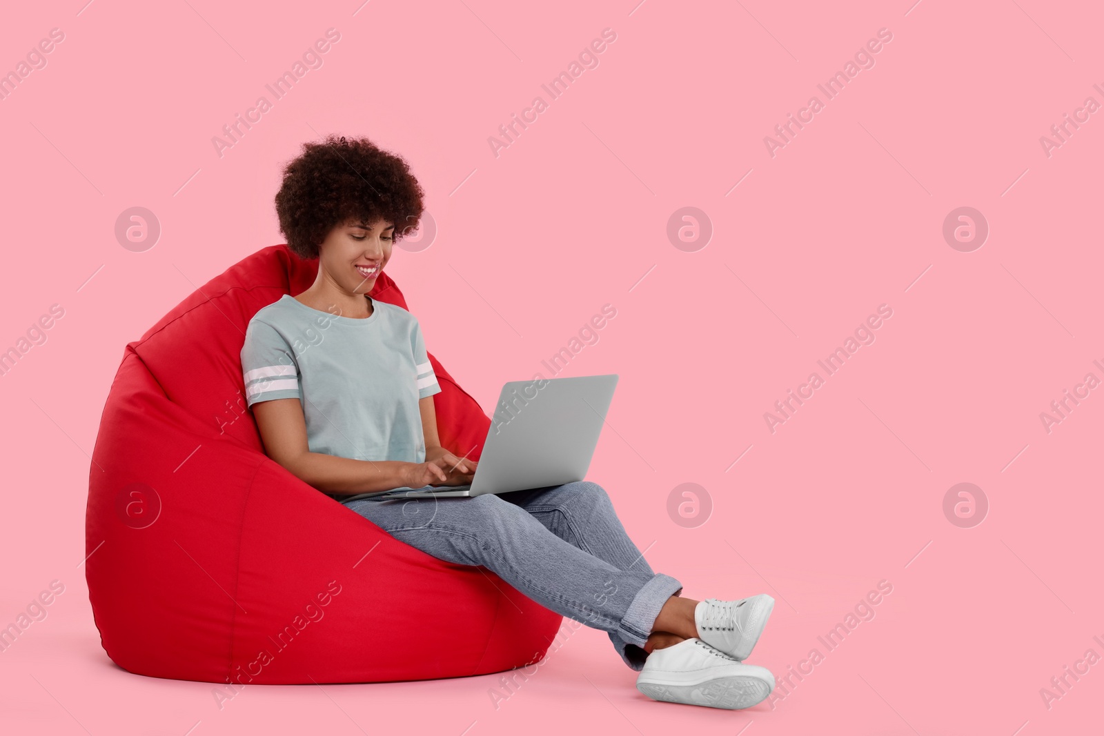 Photo of Beautiful young woman with laptop sitting on beanbag chair against pink background. Space for text