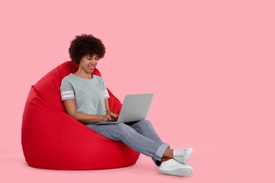 Beautiful young woman with laptop sitting on beanbag chair against pink background. Space for text