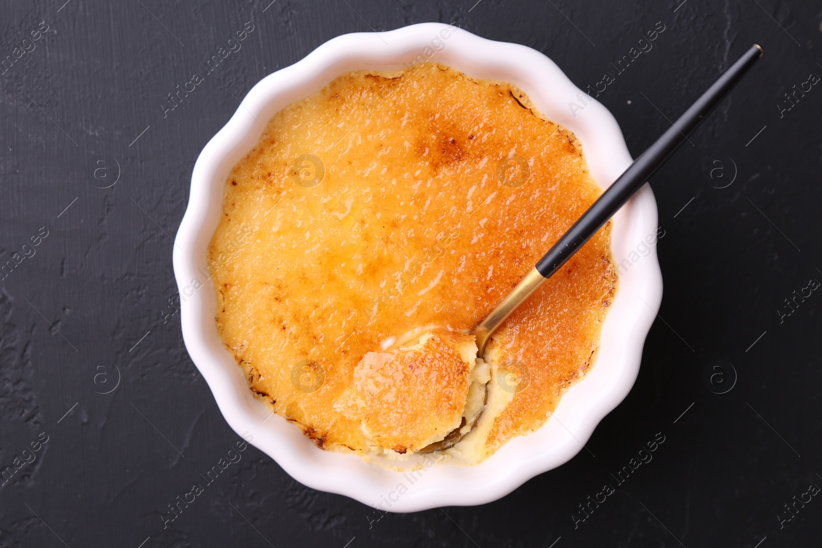 Photo of Delicious creme brulee in bowl and spoon on dark gray textured table, top view