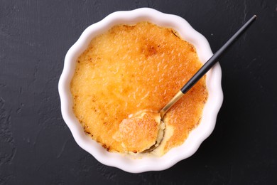 Photo of Delicious creme brulee in bowl and spoon on dark gray textured table, top view