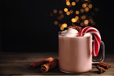 Photo of Glass cup of tasty cocoa with marshmallows and Christmas candy cane on wooden table against blurred festive lights. Space for text