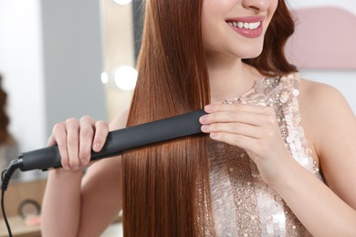 Young woman using hair iron in room, closeup