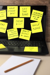 Photo of Laptop with paper notes of life-affirming phrases on beige desk indoors