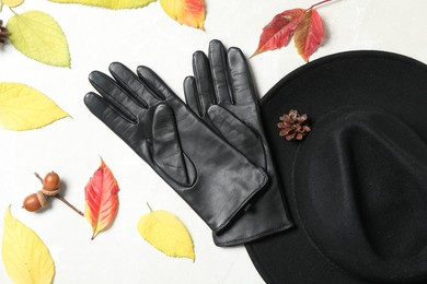 Stylish black leather gloves, hat and dry leaves on white table, flat lay