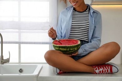Photo of Young African American woman with half of watermelon sitting on countertop in kitchen, closeup