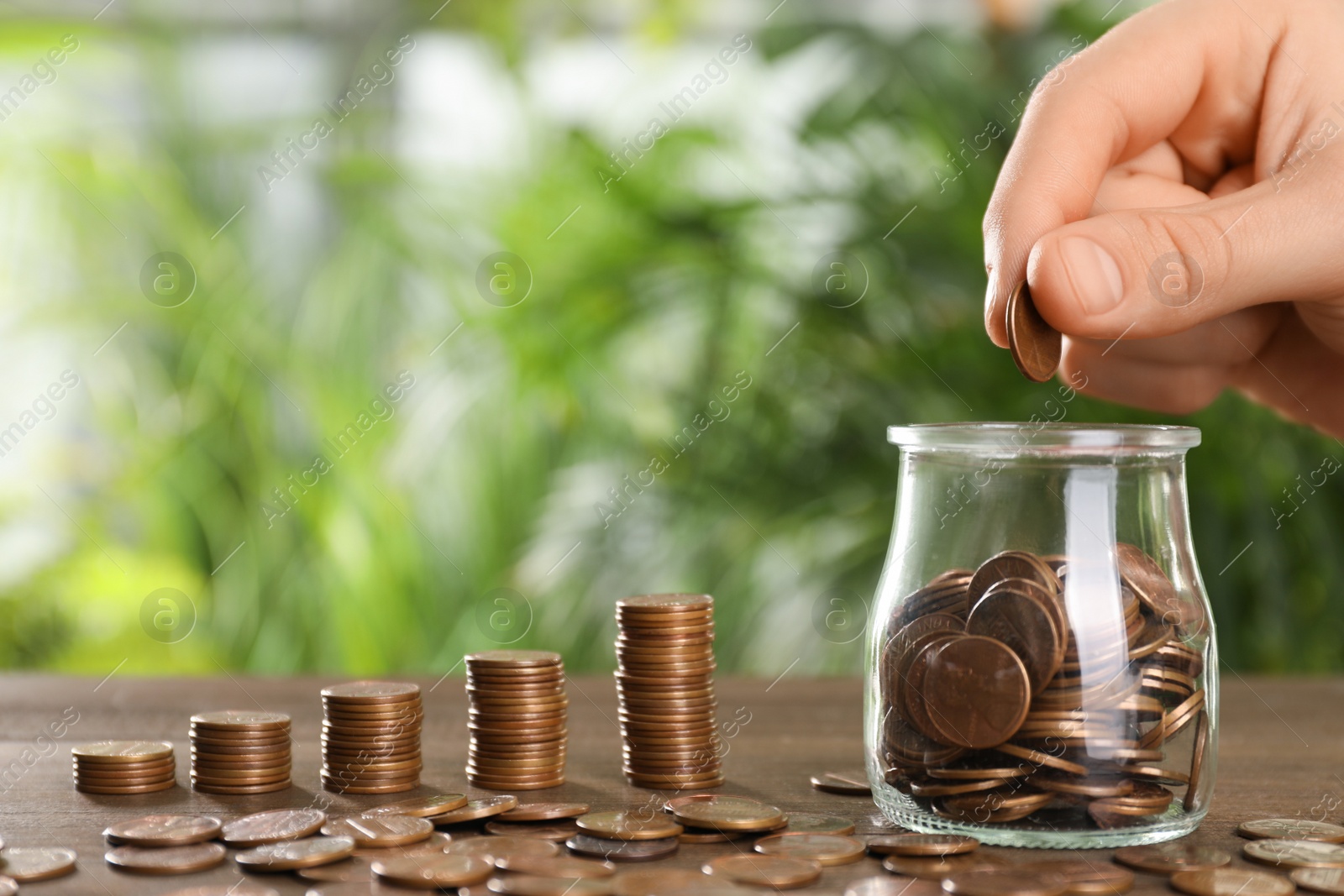 Photo of Woman putting coin into glass jar at wooden table against blurred green background, closeup. Space for text