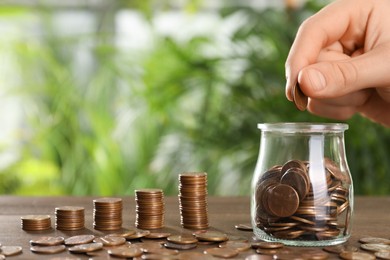 Woman putting coin into glass jar at wooden table against blurred green background, closeup. Space for text