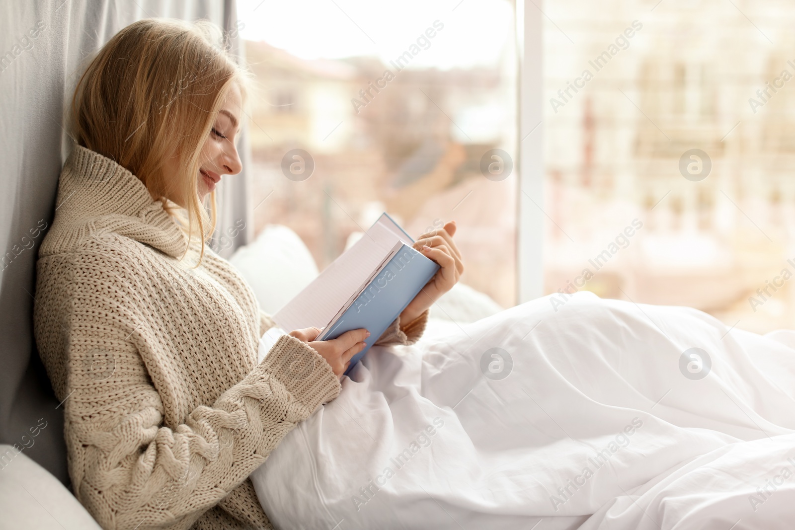 Photo of Beautiful young woman in knitted sweater sitting and reading book near window at home. Winter atmosphere