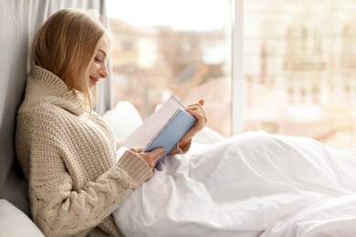 Photo of Beautiful young woman in knitted sweater sitting and reading book near window at home. Winter atmosphere