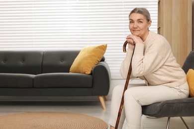 Senior woman with walking cane sitting on armchair at home. Space for text