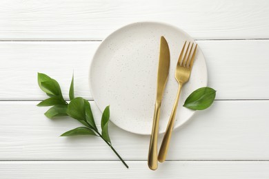 Stylish setting with cutlery, green leaves and plate on white wooden table, flat lay