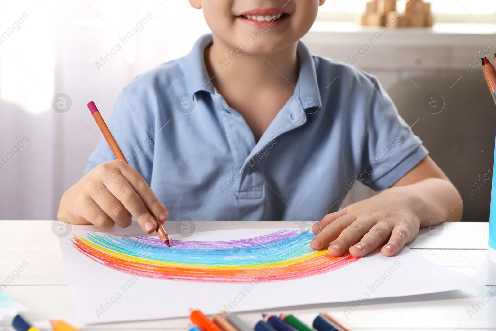 Photo of Little boy drawing rainbow with pencil at white wooden table indoors, closeup. Child`s art