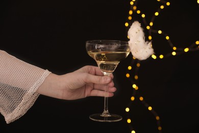 Photo of Woman holding glass of cocktail decorated with tasty cotton candy on black background with blurred lights, closeup