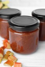 Photo of Jars of tasty rhubarb jam and cut stalks on white wooden table, closeup