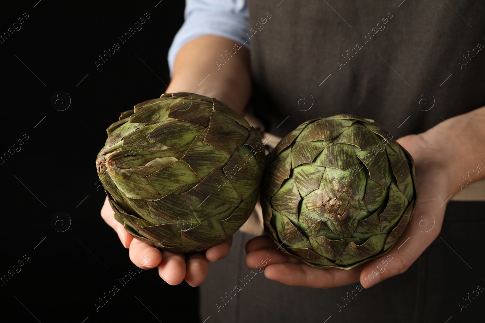 Photo of Woman holding fresh raw artichokes on black background, closeup