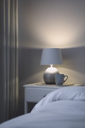 Stylish lamp and cup of drink on white nightstand in bedroom