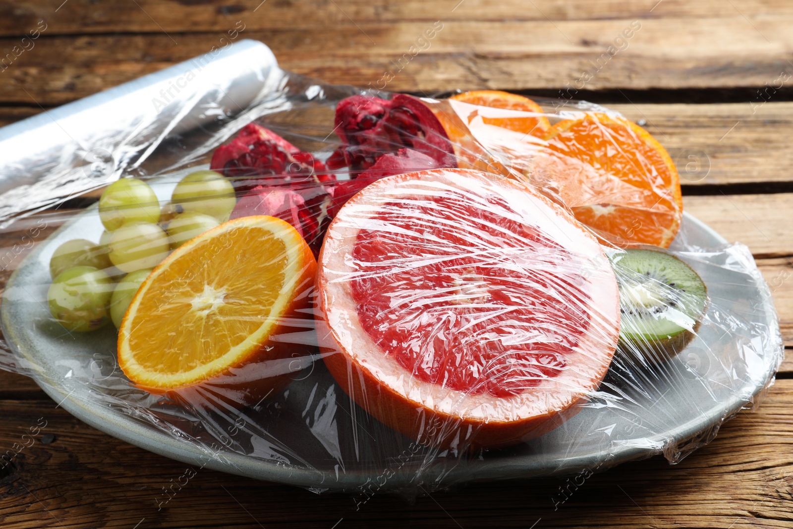 Photo of Plate of fresh fruits with plastic food wrap on wooden table, closeup