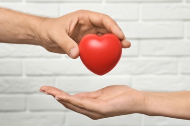 Man giving red heart to woman at white brick wall, closeup. Donation concept