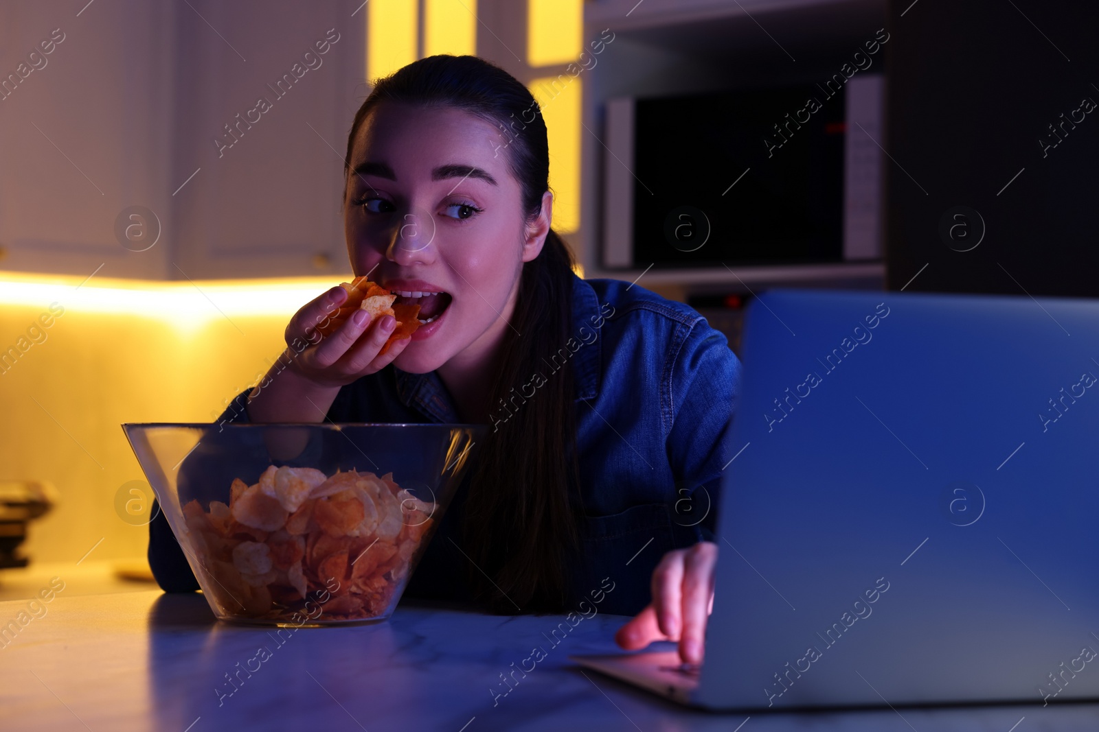 Photo of Young woman eating chips while using laptop in kitchen at night. Bad habit