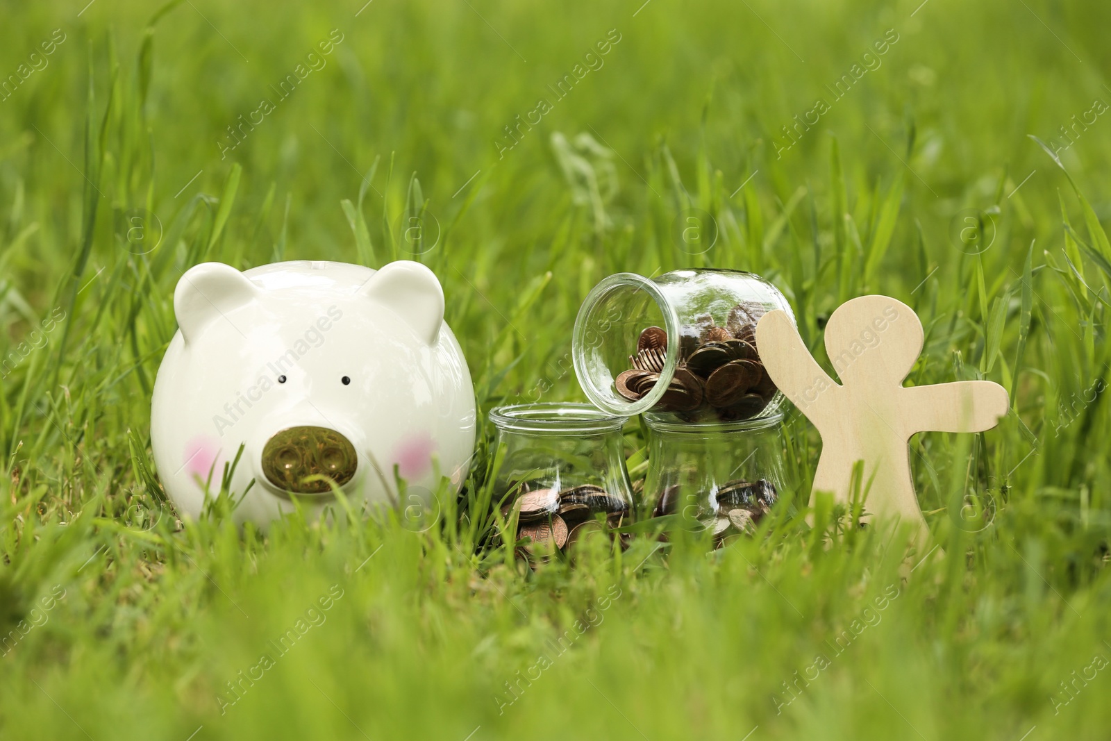 Photo of Cute piggy bank, wooden person and jars with coins on green grass in park