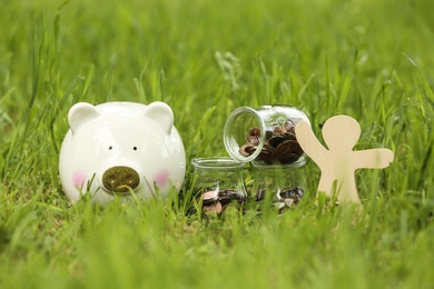 Photo of Cute piggy bank, wooden person and jars with coins on green grass in park