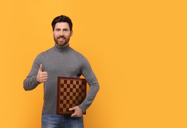 Photo of Smiling man holding chessboard and showing thumb up on yellow background. Space for text