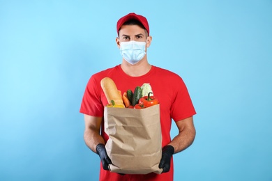 Photo of Courier in medical mask holding paper bag with food on light blue background. Delivery service during quarantine due to Covid-19 outbreak
