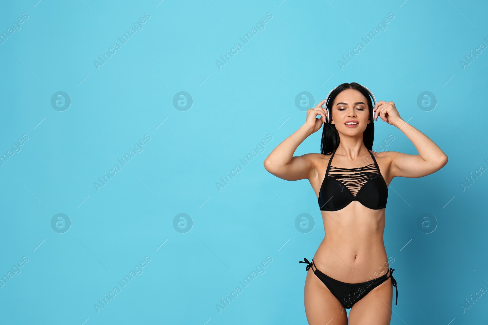 Photo of Beautiful young woman in black bikini with headphones on light blue background. Space for text