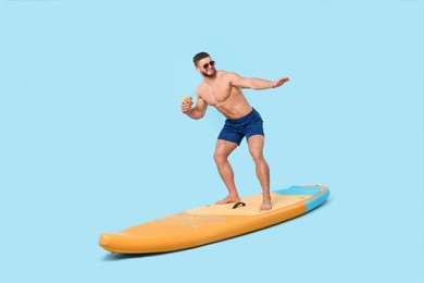 Happy man with refreshing drink balancing on SUP board against light blue background