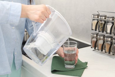 Photo of Woman pouring water from filter jug into glass in kitchen, closeup