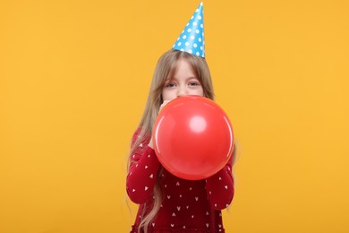 Cute little girl in party hat inflating balloon on yellow background