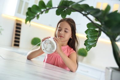 Photo of Little girl enjoying air flow from portable fan at home. Summer heat