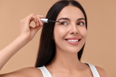 Beautiful woman applying serum onto her eyelashes on beige background. Cosmetic product