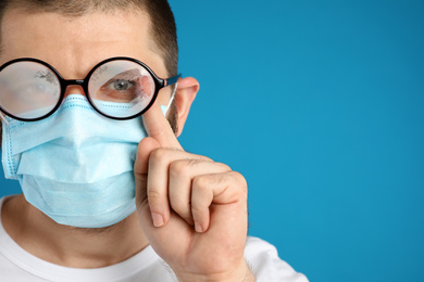 Photo of Man wiping foggy glasses caused by wearing disposable mask on blue background, space for text. Protective measure during coronavirus pandemic