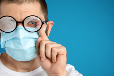 Man wiping foggy glasses caused by wearing disposable mask on blue background, space for text. Protective measure during coronavirus pandemic