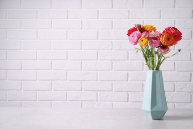 Beautiful fresh ranunculus flowers in vase on white table near brick wall, space for text
