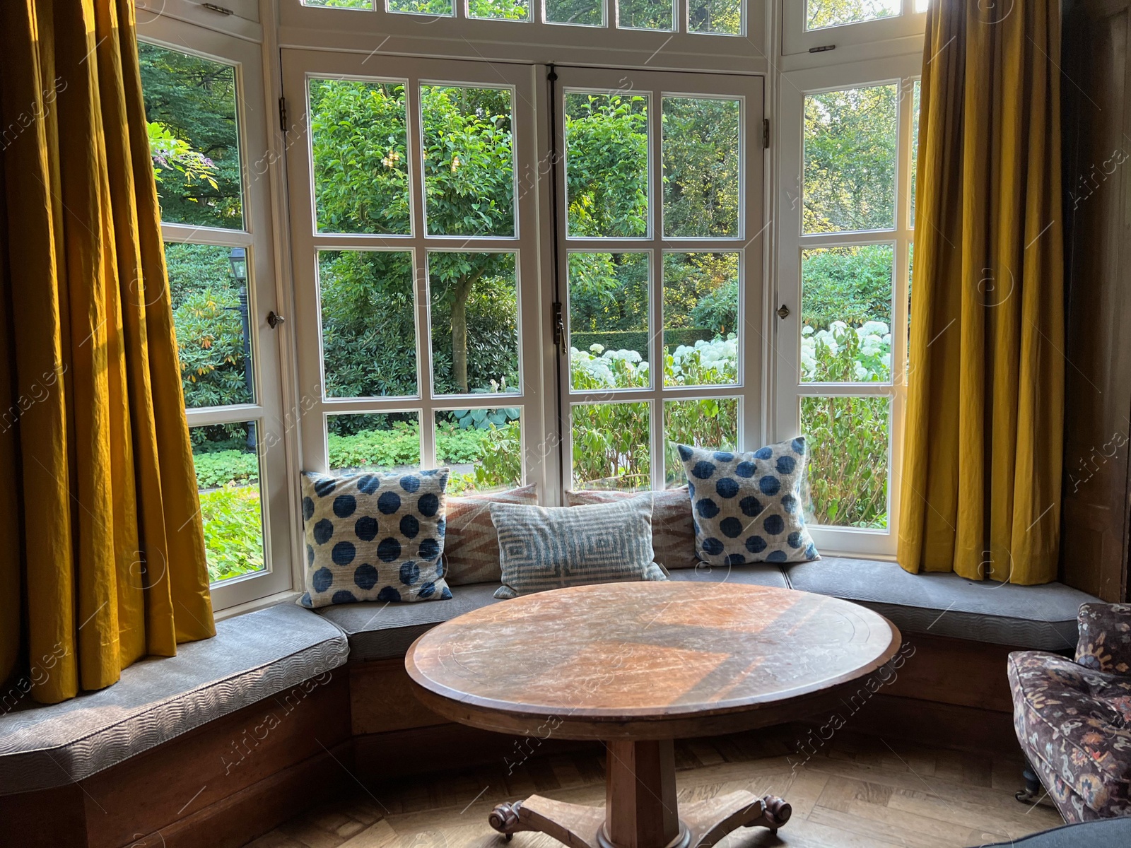 Photo of Wooden table near cozy bay window seat with cushions in room. Interior design