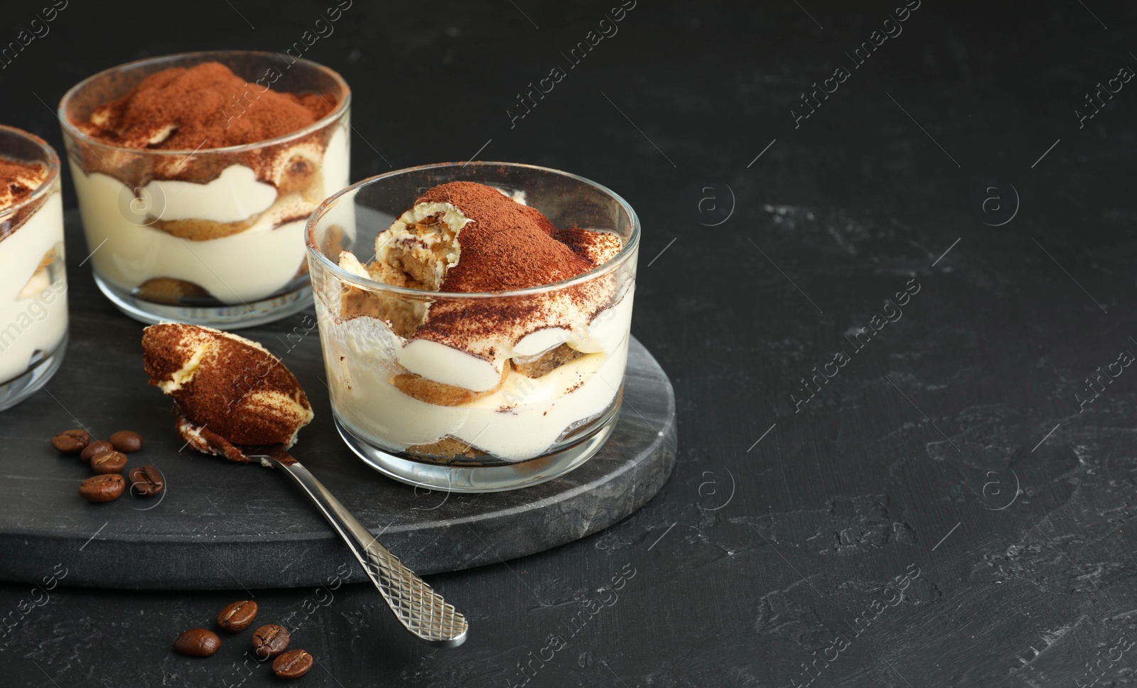 Photo of Delicious tiramisu in glasses, spoon and coffee beans on black table, space for text