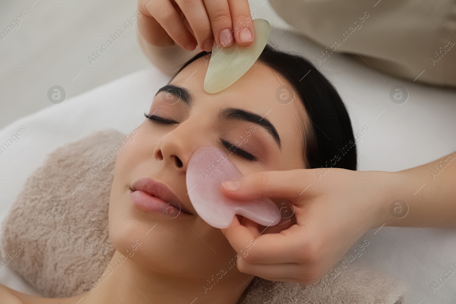 Photo of Young woman receiving facial massage with gua sha tools in beauty salon