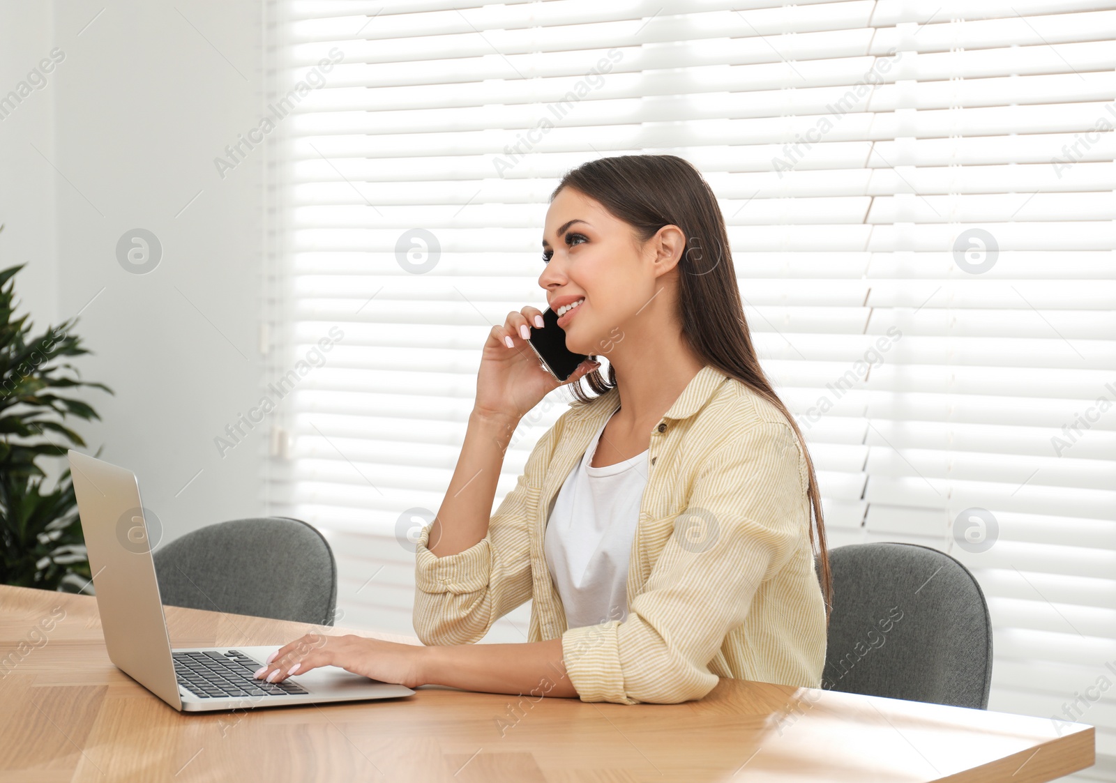 Photo of Young woman talking on phone while working with laptop in office