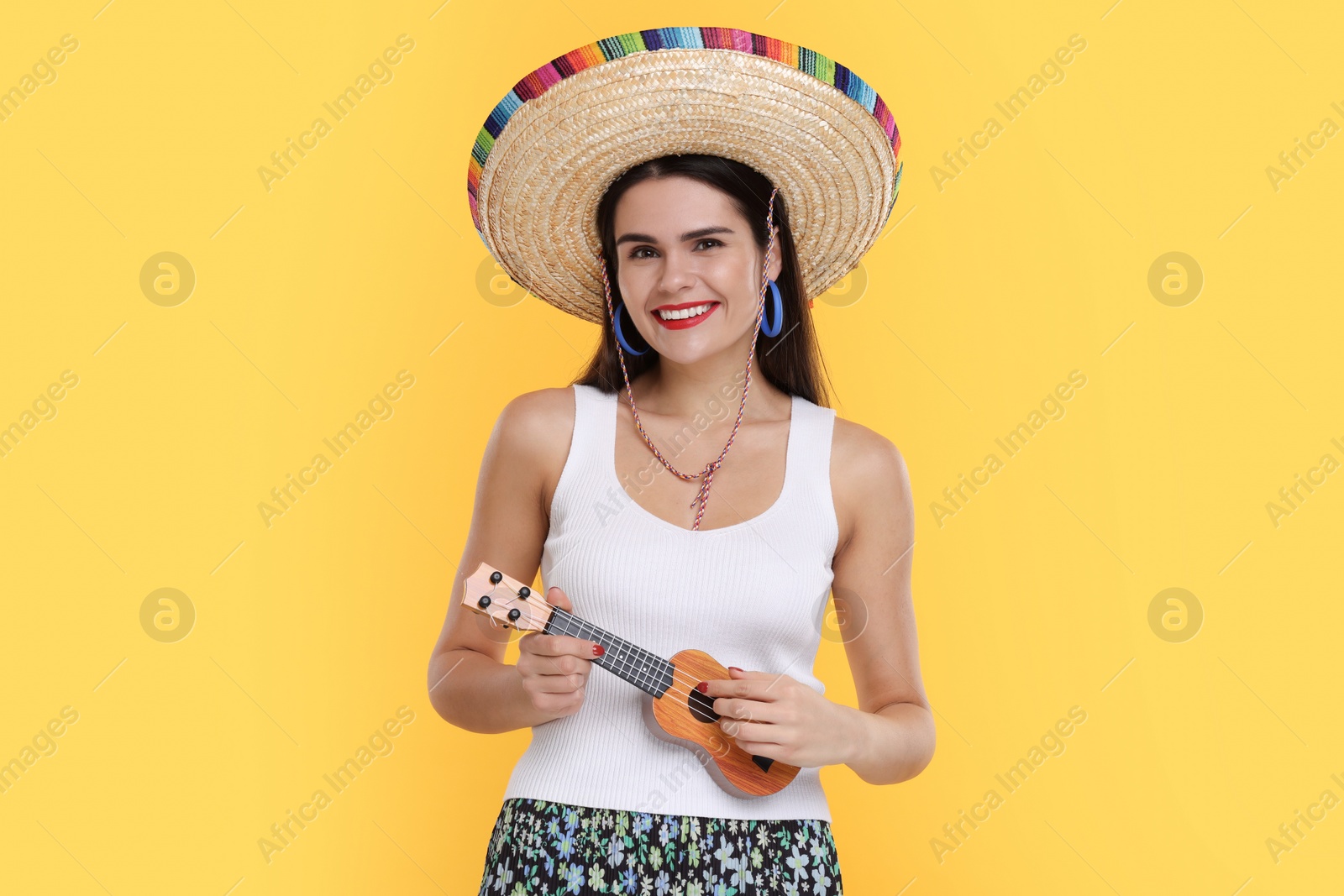 Photo of Young woman in Mexican sombrero hat playing ukulele on yellow background