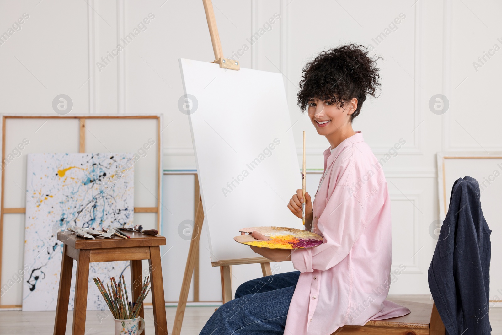 Photo of Young woman holding brush and artist`s palette near easel with canvas in studio